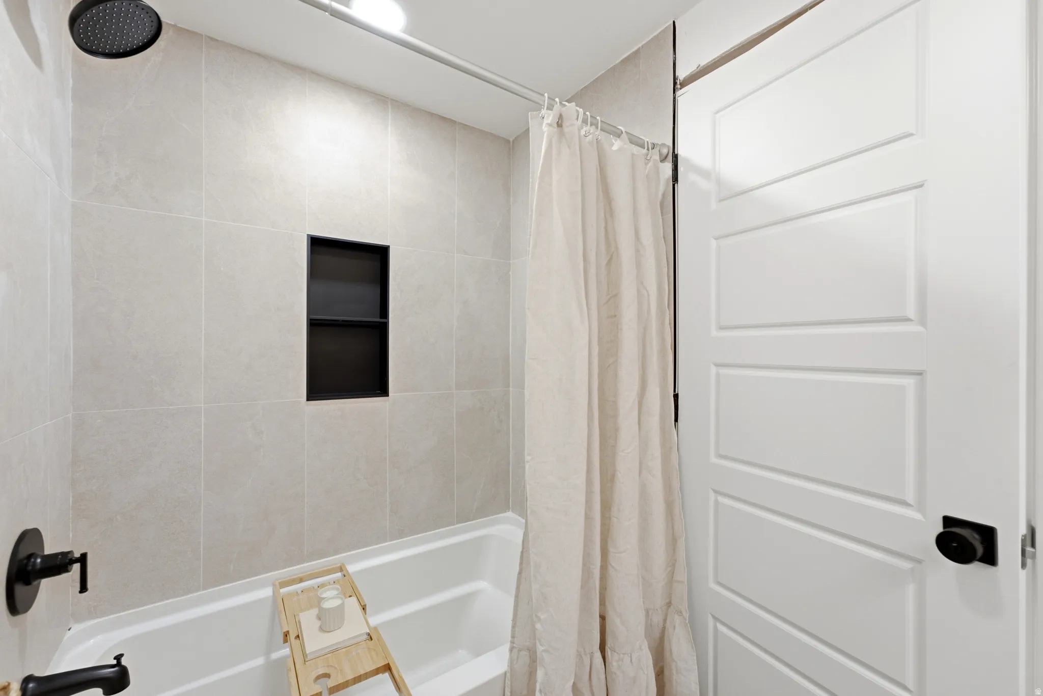 Bathtub and shower combination featuring neutral-toned wall tiling, a built-in shower niche with dark shelving, matte black fixtures, and a ceiling-mounted showerhead