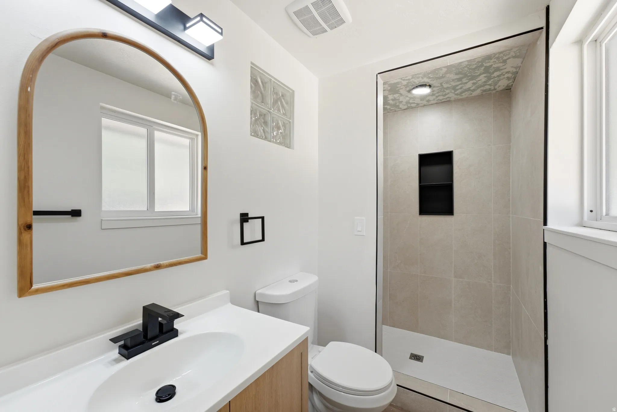 Bathroom featuring a light wood-finish vanity with a white countertop and oval sink, matte black faucet, and an arched wood-frame mirror
