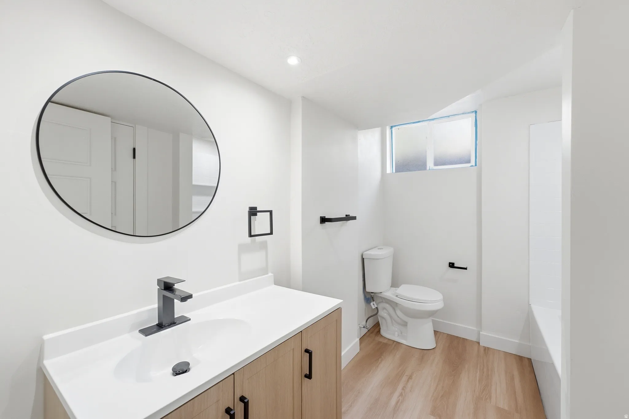 Bathroom featuring a wood-finish vanity with a white countertop and integrated sink, matte black faucet, and a circular black-framed mirror