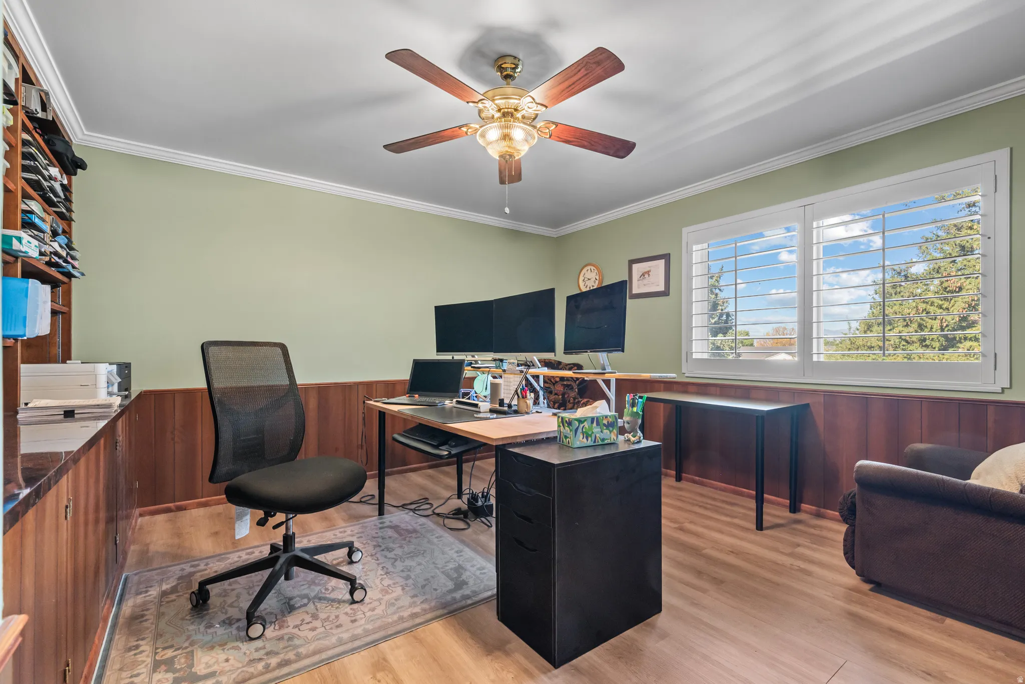 Room featuring wood-finish flooring, a ceiling fan with integrated lighting, crown molding, and a large window with white plantation shutters