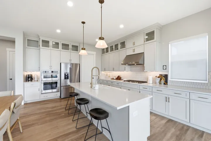Gourmet kitchen featuring a central island with countertop seating, light wood-finish flooring, white shaker cabinetry with glass upper doors, stainless steel appliances, and a white geometric tile backsplash