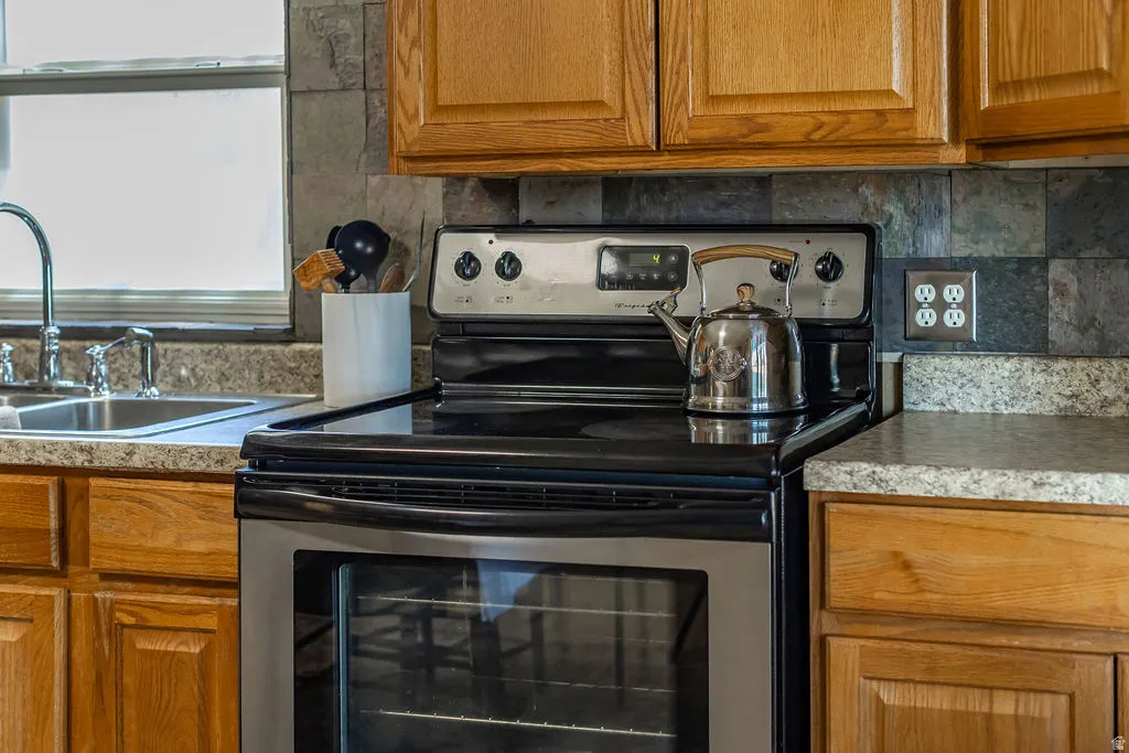 Stainless steel electric range with a visible oven, slate-style tile backsplash, wood-finish cabinetry, and speckled countertops