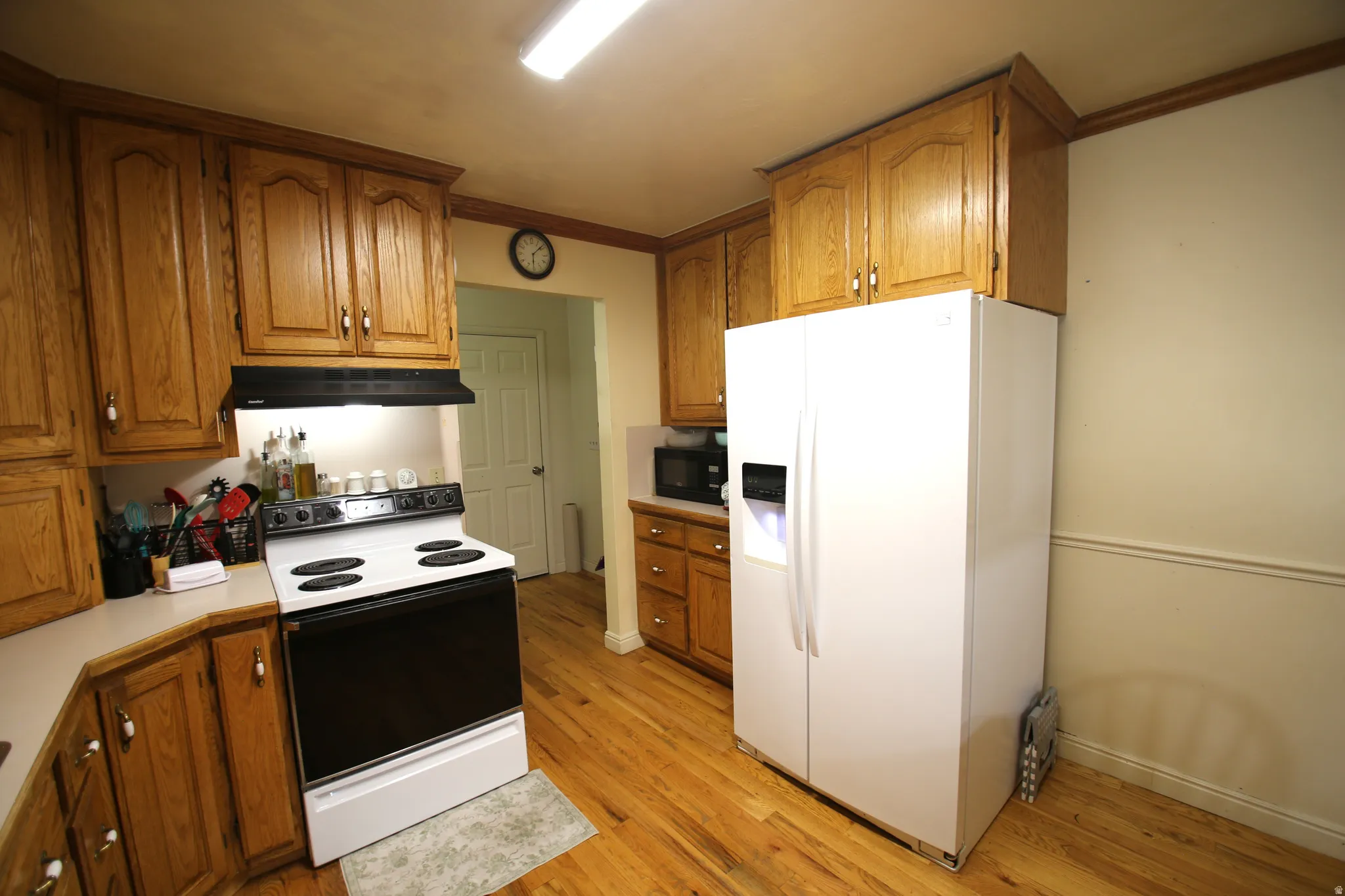 Kitchen featuring wood-finish flooring, wood cabinetry with upper and lower storage, white appliances including a side-by-side refrigerator, and a white electric range with an overhead vent hood