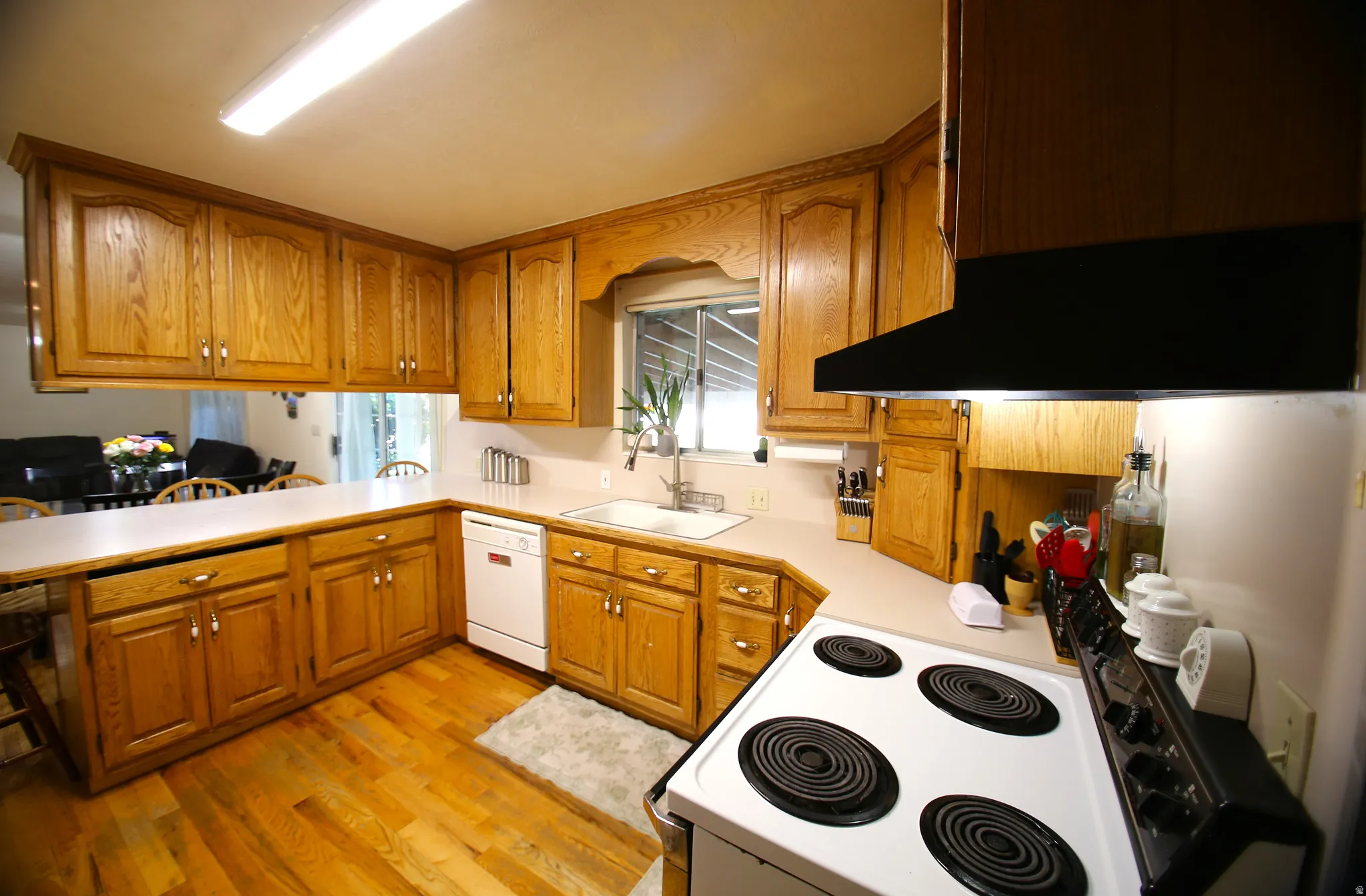 Kitchen featuring wood-finish flooring, wood cabinetry with upper and lower storage, a white built-in dishwasher, white countertops, and a white built-in sink with a window above