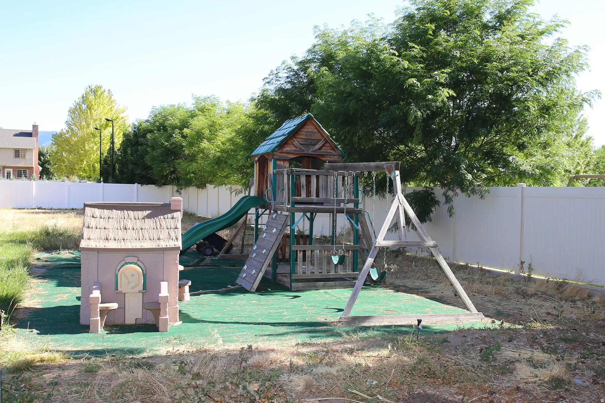 Spacious backyard featuring a wooden play structure with slide, a separate playhouse, and a swing set, all surrounded by a white privacy fence