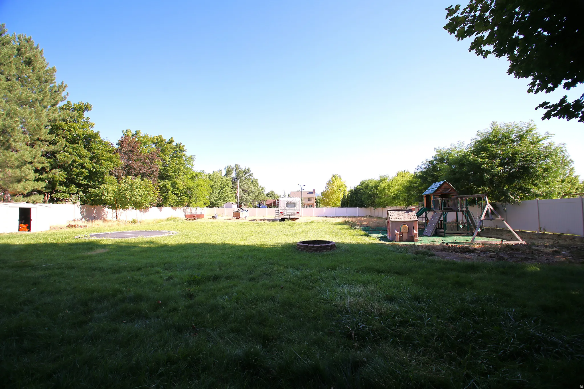 Expansive backyard featuring a large green space, privacy fencing, mature trees, and a built-in fire pit