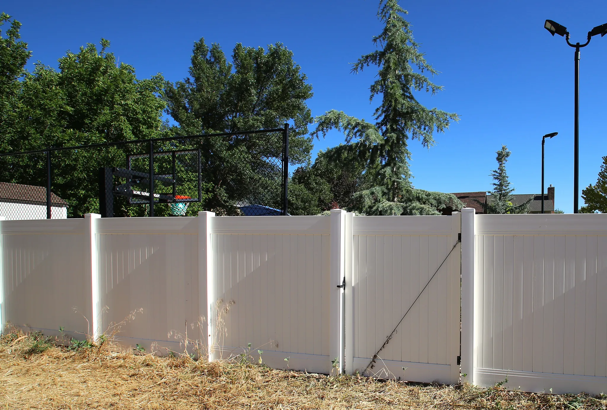 Outdoor area featuring a white privacy fence with a functional gate, complemented by mature perimeter trees and a visible basketball hoop structure beyond the fence