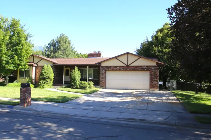 Brick ranch-style home featuring a two-car garage, mature landscaping, a covered front porch, and a paved driveway