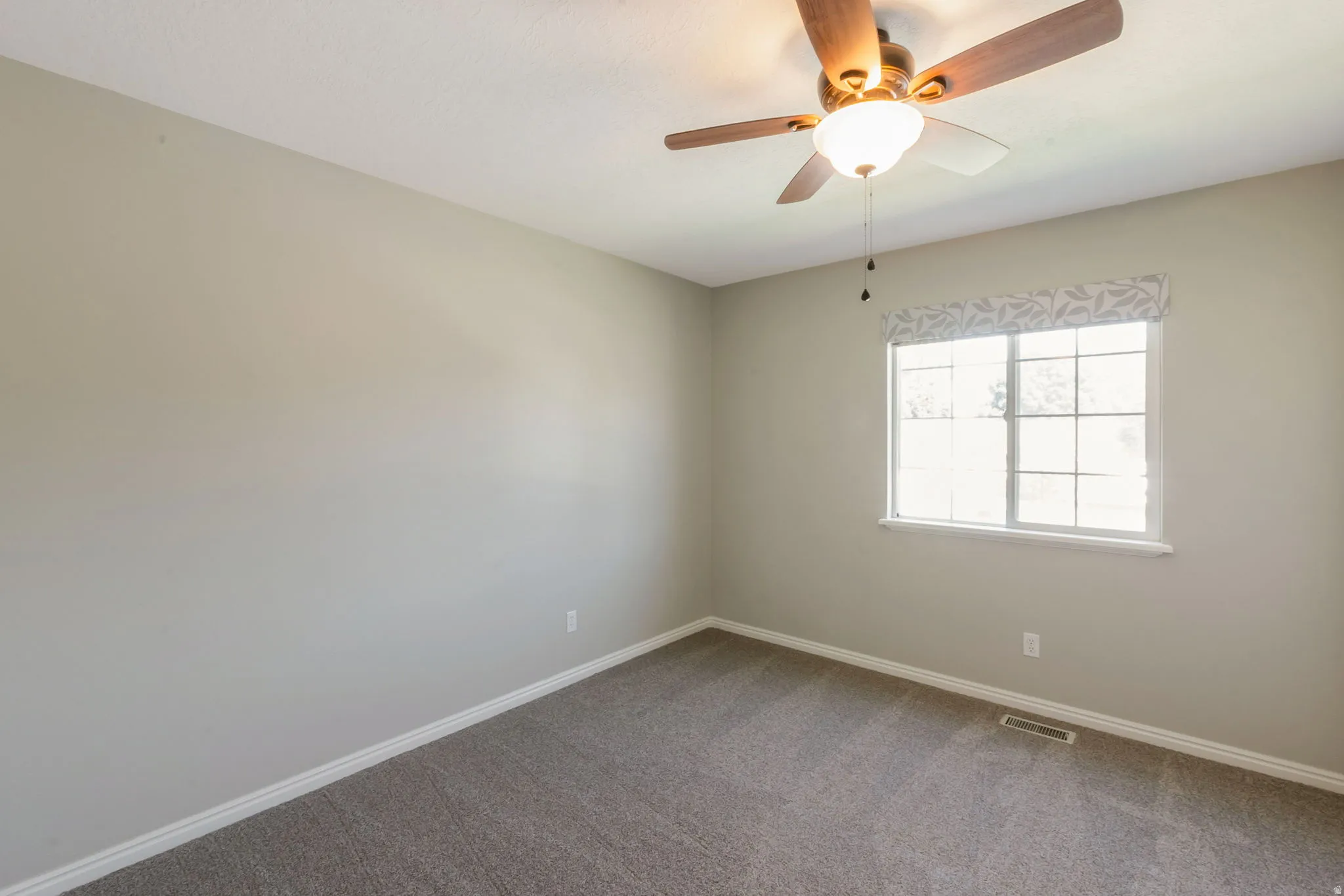Neutral-toned room featuring a large window with a decorative valance, a ceiling fan with integrated lighting, and gray carpet flooring