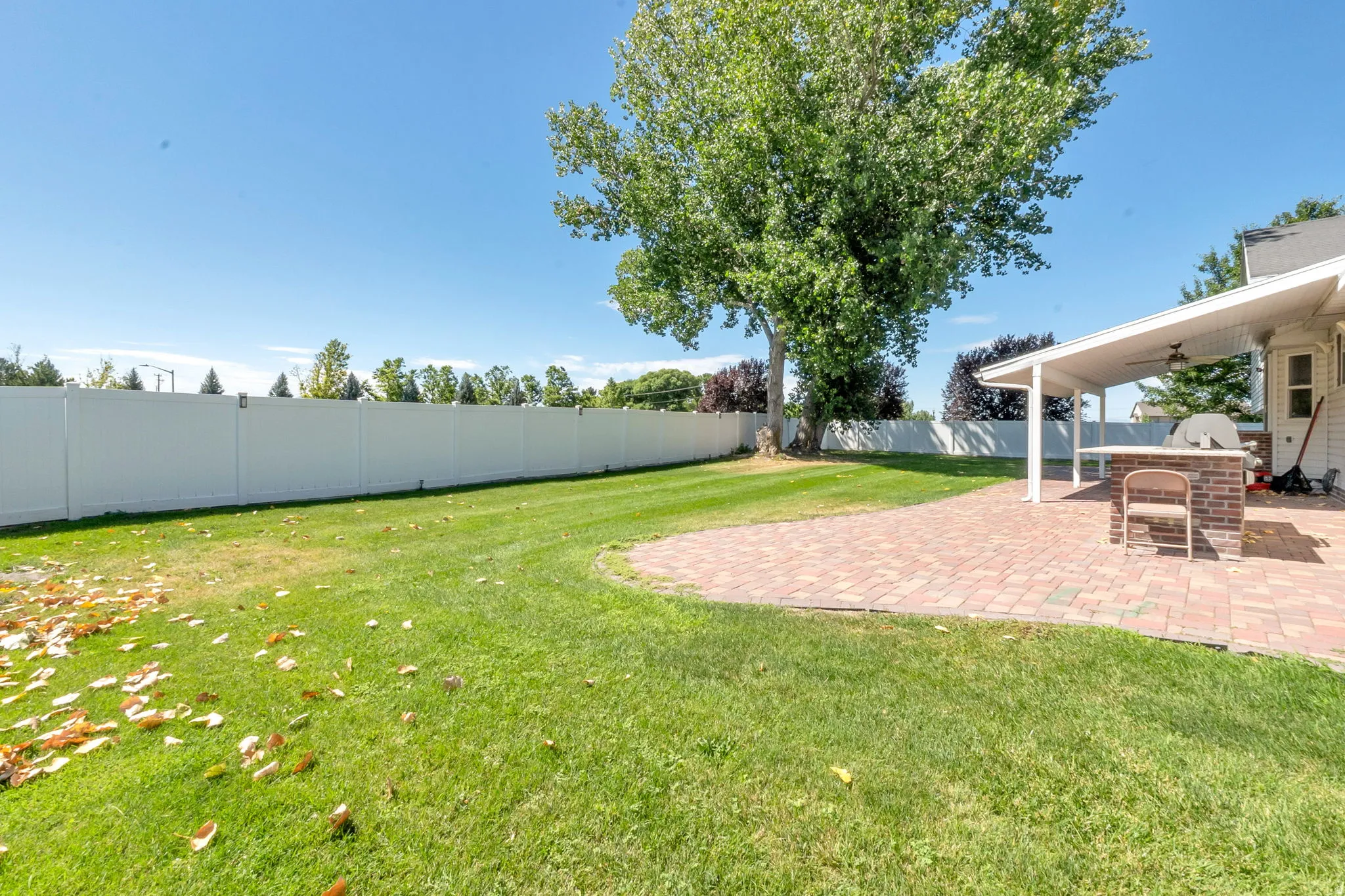 Spacious backyard featuring a large brick paver patio with a white covered pergola, expansive lawn, mature shade tree, and a white privacy fence