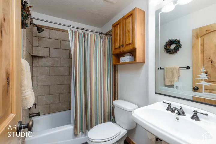 Full bathroom featuring a tub and shower combination with stone-look tile surround, a wall-mounted sink with a dark bronze-finish faucet, a toilet, a wood-finish wall cabinet, and a mirrored vanity with a decorative etched design