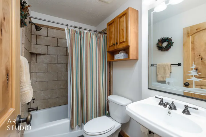 Full bathroom featuring a tub and shower combination with stone-look tile surround, a wall-mounted sink with a dark bronze-finish faucet, a toilet, a wood-finish wall cabinet, and a mirrored vanity with a decorative etched design