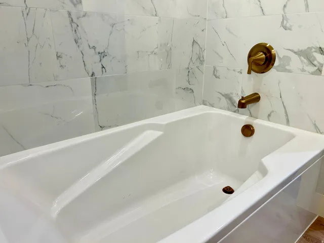 Bathtub featuring a white finish, bronze fixtures, and surrounding marble-look wall tiling
