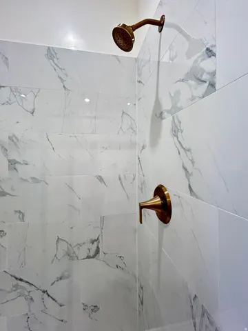 Shower enclosure featuring large format white tiled walls with gray veining, a bronze-finish shower head, and coordinating bronze-finish control handle