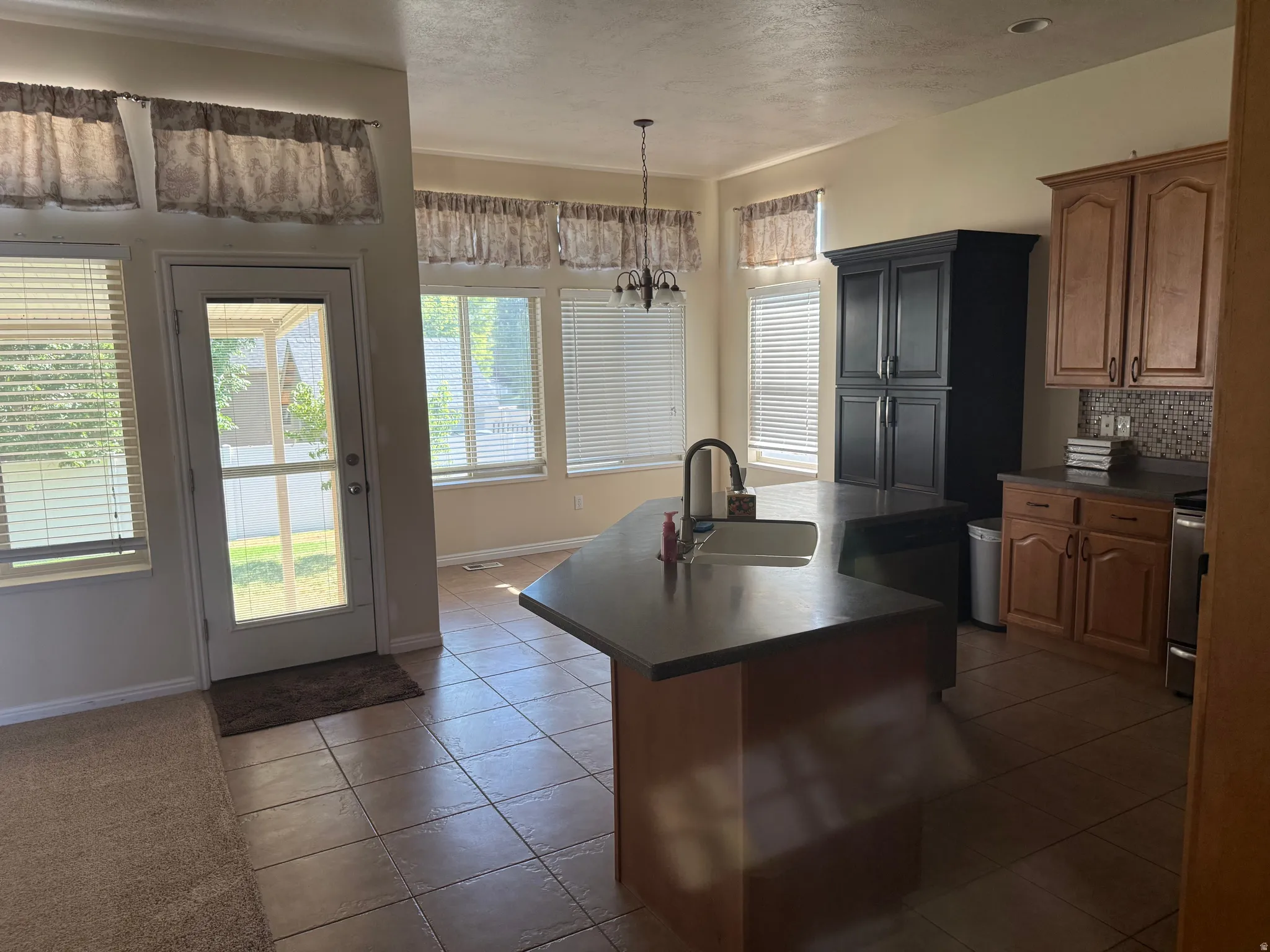 Expansive kitchen featuring a central island with an integrated sink, wood-finish cabinetry, a black pantry cabinet, and tile flooring