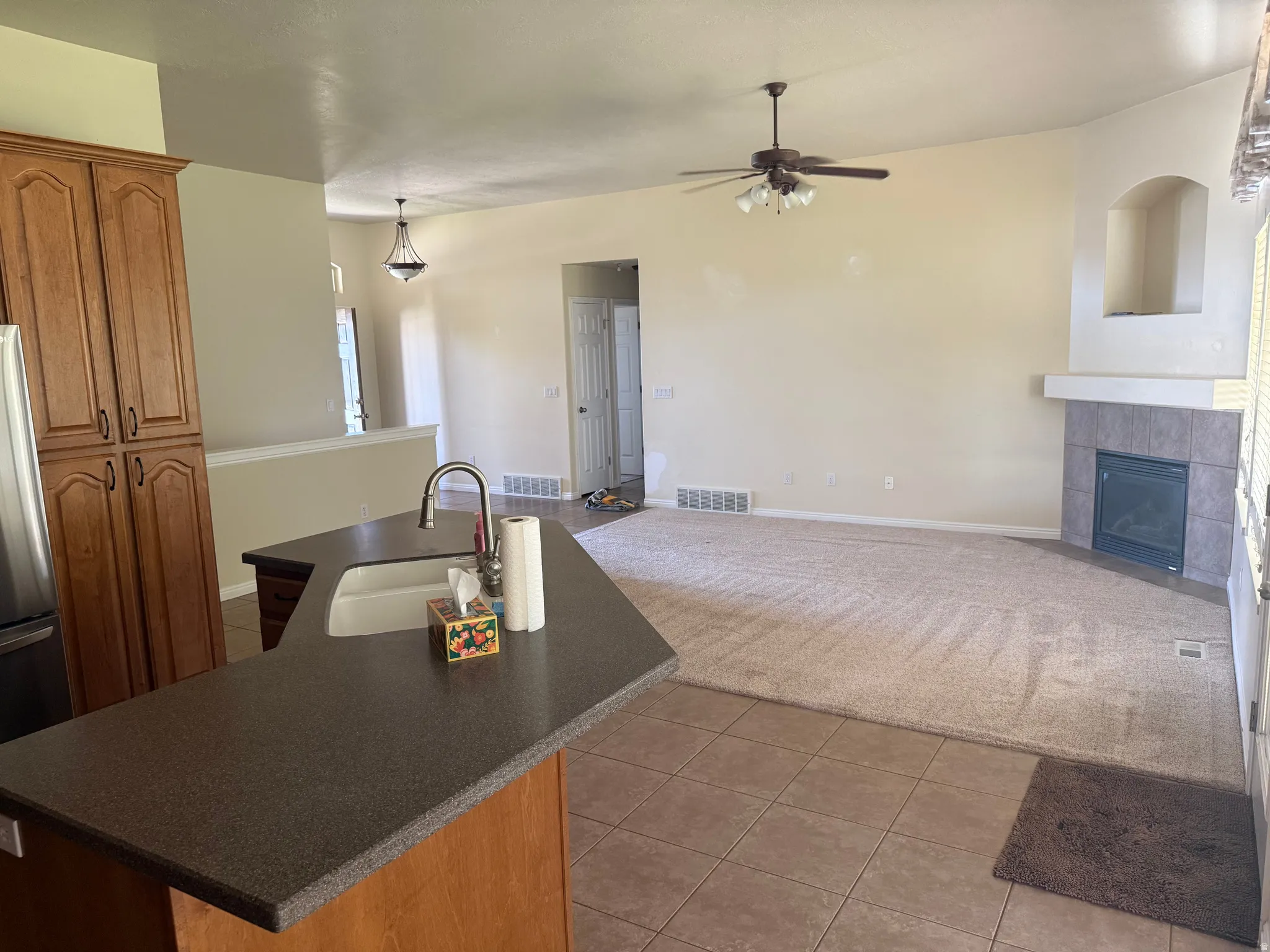 Open-concept living area featuring a kitchen island with a double basin sink, ample wood cabinetry, a ceiling fan with integrated lighting, a gas fireplace with a tiled surround, and a decorative arched niche