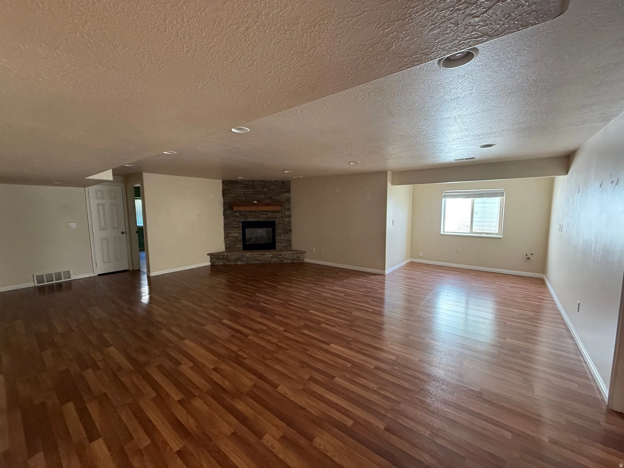 Expansive room featuring wood-finish flooring, a corner fireplace with stone surround and a wooden mantel, recessed lighting, and a window with blinds