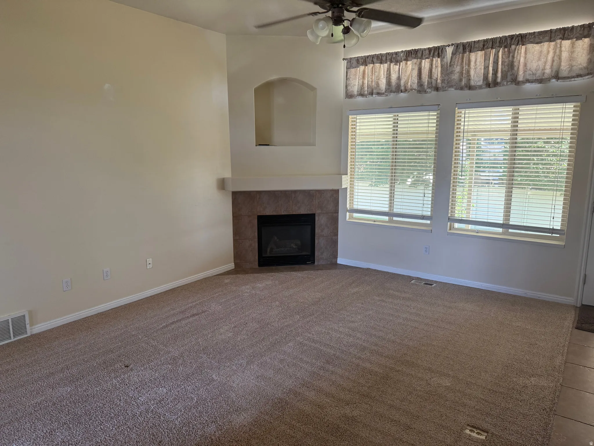 Living area featuring an integrated fireplace with a tile surround and mantel, two large windows with horizontal blinds, a ceiling fan with integrated lighting, and a neutral carpet