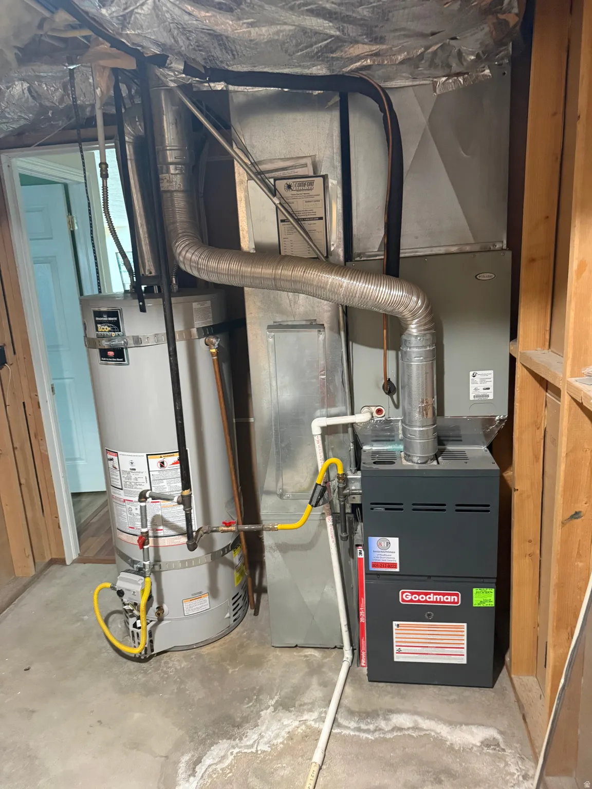 Goodman brand furnace and water heater installation with visible ductwork, gas lines, and PVC piping on a concrete floor