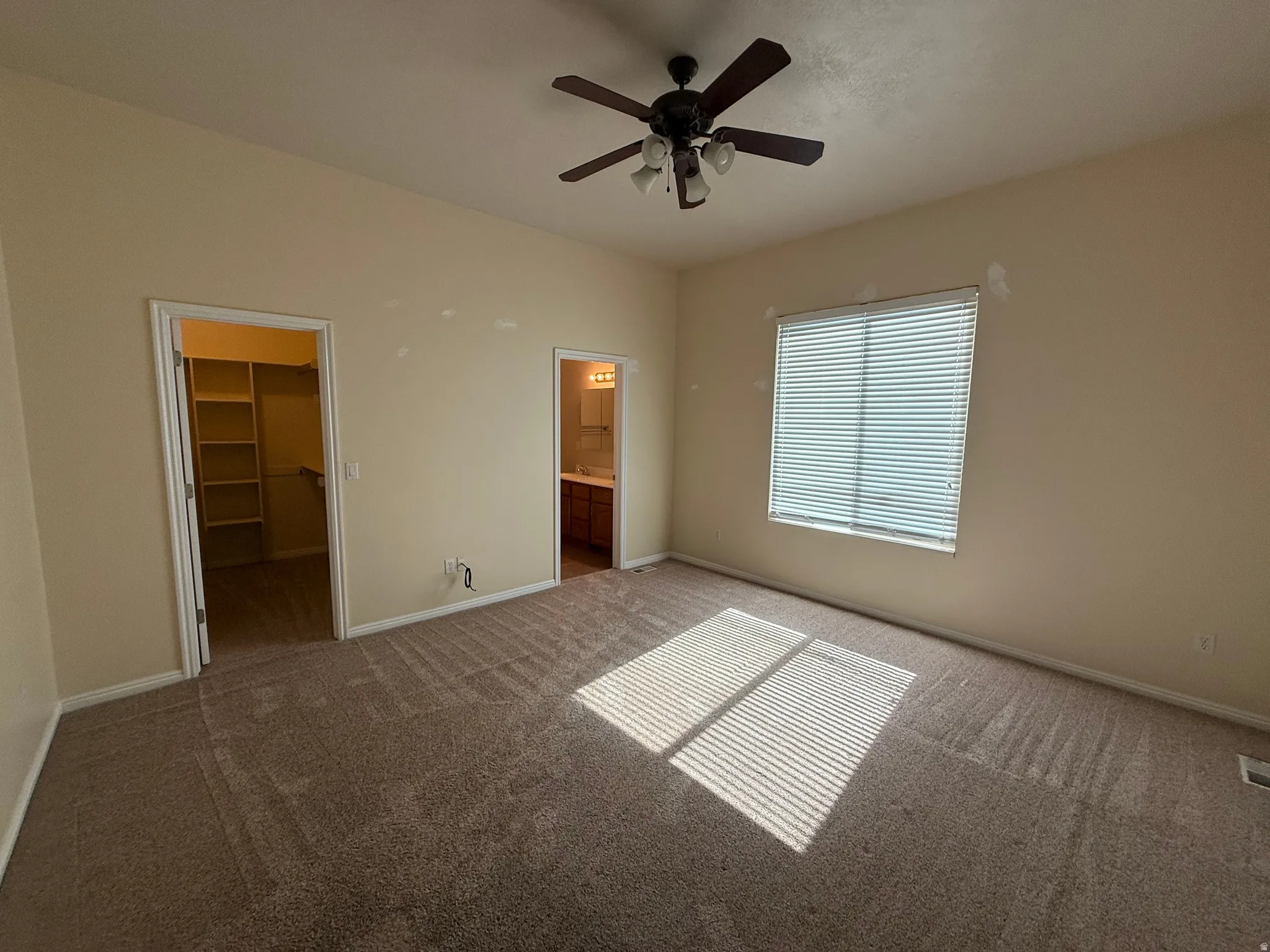 Carpeted room featuring a ceiling fan with integrated lighting, a window with horizontal blinds, and two doorways leading to a walk-in closet with built-in shelving and a bathroom with a vanity and mirror
