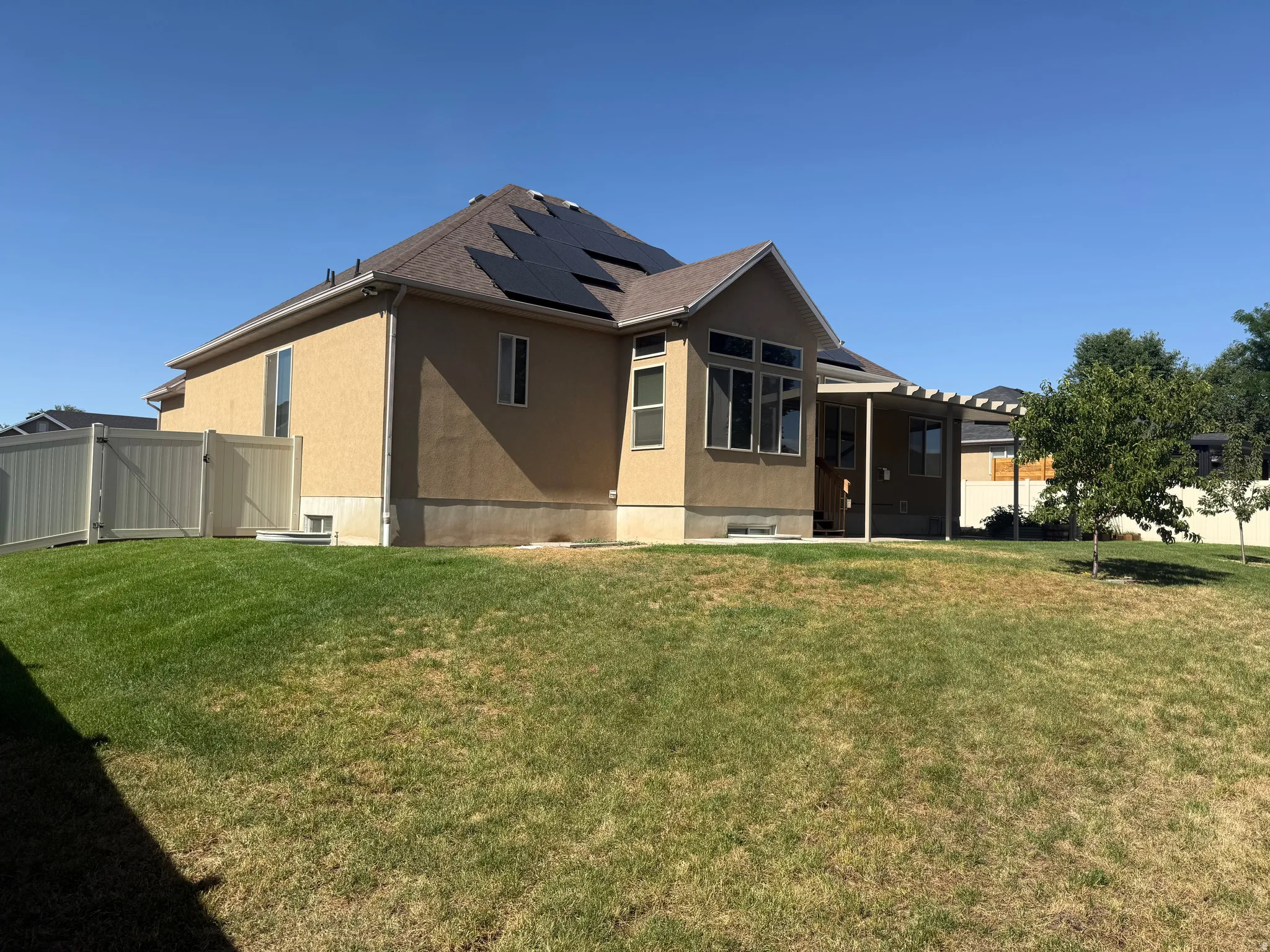 Expansive rear yard featuring a stucco exterior, multiple solar panels, a covered patio, vinyl fencing, and mature trees