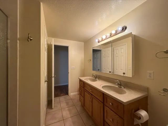 Dual sink vanity with wood-finish cabinetry, light-toned countertop, wall-mounted light fixture, and tiled flooring
