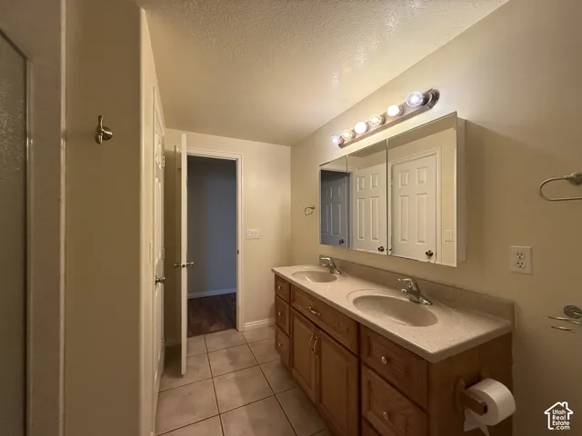 Dual sink vanity with wood-finish cabinetry, light-toned countertop, wall-mounted light fixture, and tiled flooring