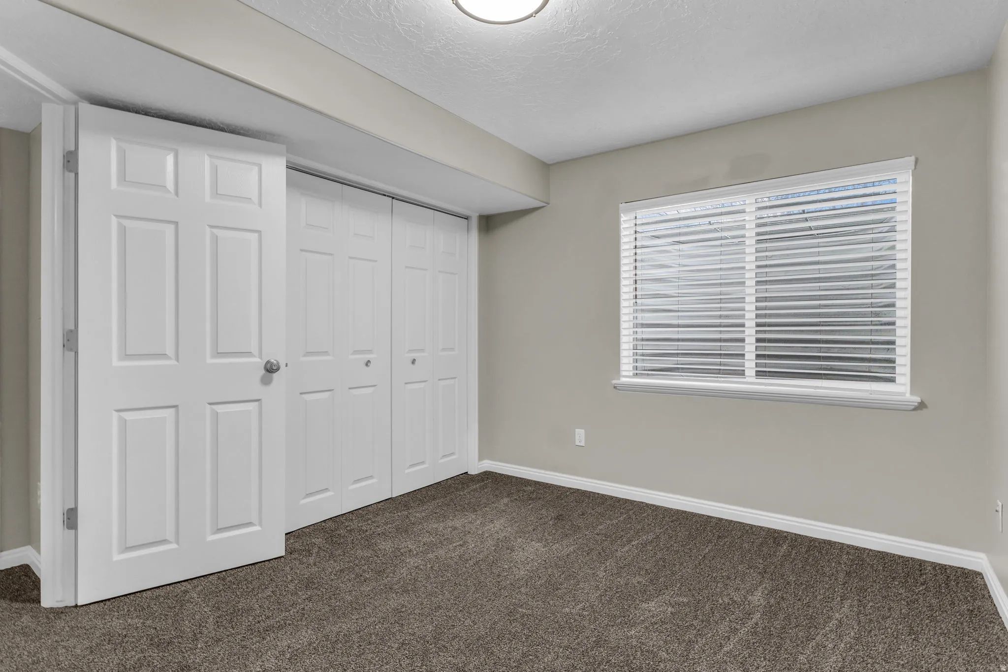 Carpeted room featuring a wide window with blinds, bi-fold closet doors, a white paneled door, and neutral wall paint