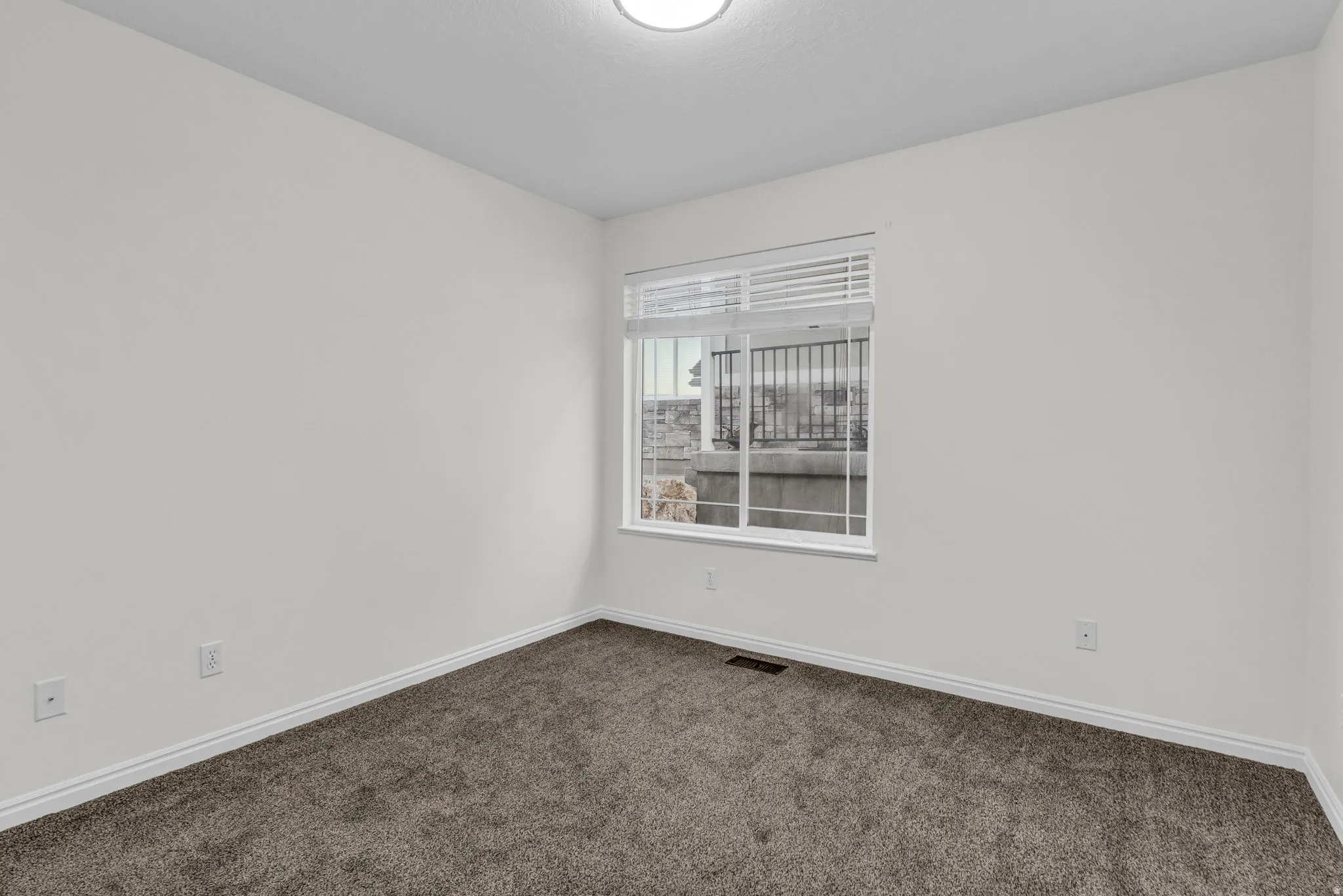 Bright interior room featuring a window with horizontal blinds, plush dark gray carpeting, crisp white baseboards, and a flush-mount ceiling light fixture
