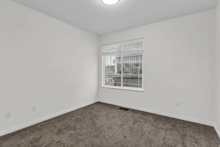 Bright interior room featuring a window with horizontal blinds, plush dark gray carpeting, crisp white baseboards, and a flush-mount ceiling light fixture