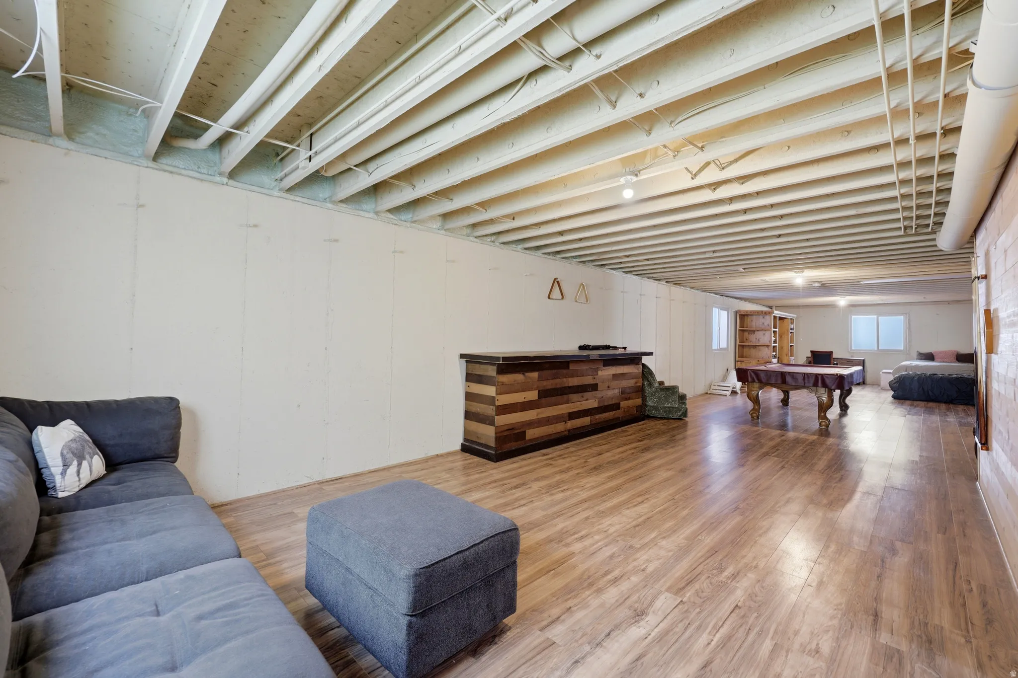 Expansive finished basement featuring wood-finish flooring, exposed beam ceiling, a multi-tone wood-paneled bar, and two windows providing natural light