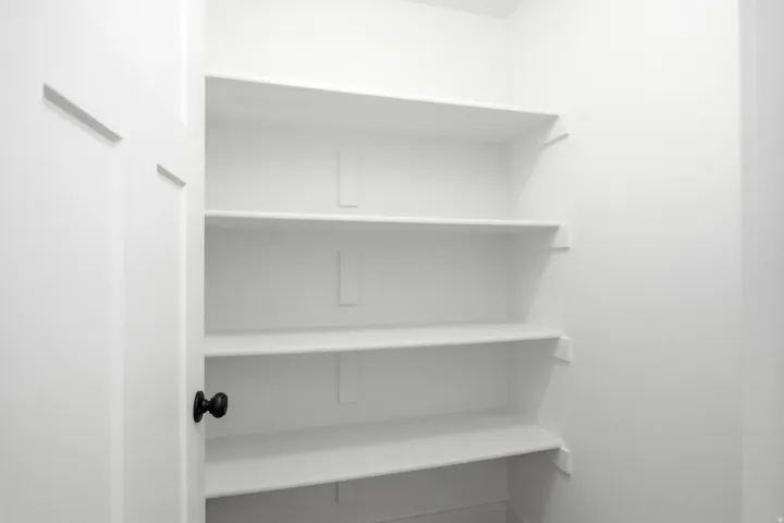 Interior pantry featuring four built-in white shelves, white walls, and a white panel door with a black knob
