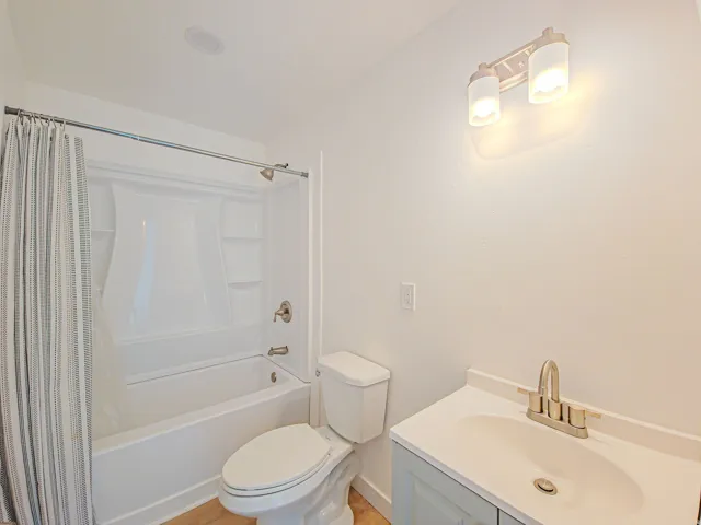 White shower-over-tub with integrated shelving, brushed nickel fixtures, light wood-finish flooring, recessed ceiling lighting, and a vanity with a white countertop and undermount sink