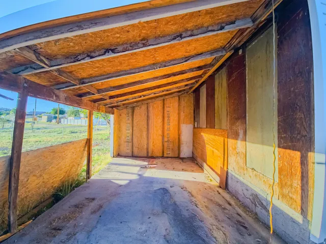 Carport structure featuring a concrete floor, plywood walls, and an exposed beam ceiling