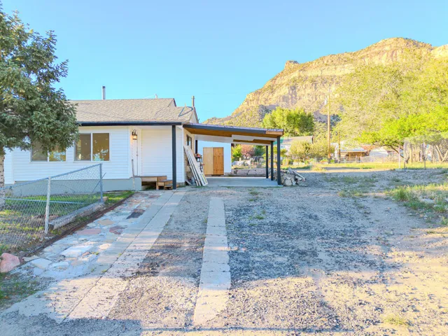 White siding exterior with a dark-pitched roof and a covered carport, storage in back. Large lot with mature trees.