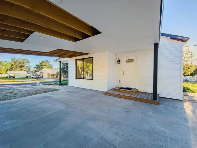 Covered concrete parking area with exposed beam ceiling detail, white siding exterior, dark framed window, and a white entry door with a decorative arch