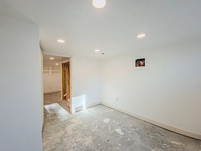 Recessed ceiling lighting illuminates a room with unfinished concrete flooring and painted white walls, new carpet on the way!