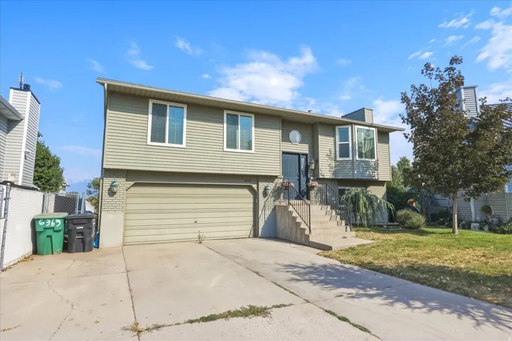Split-level residence featuring a two-car garage, light-colored siding, and a front entry accessed by concrete stairs with black metal railings