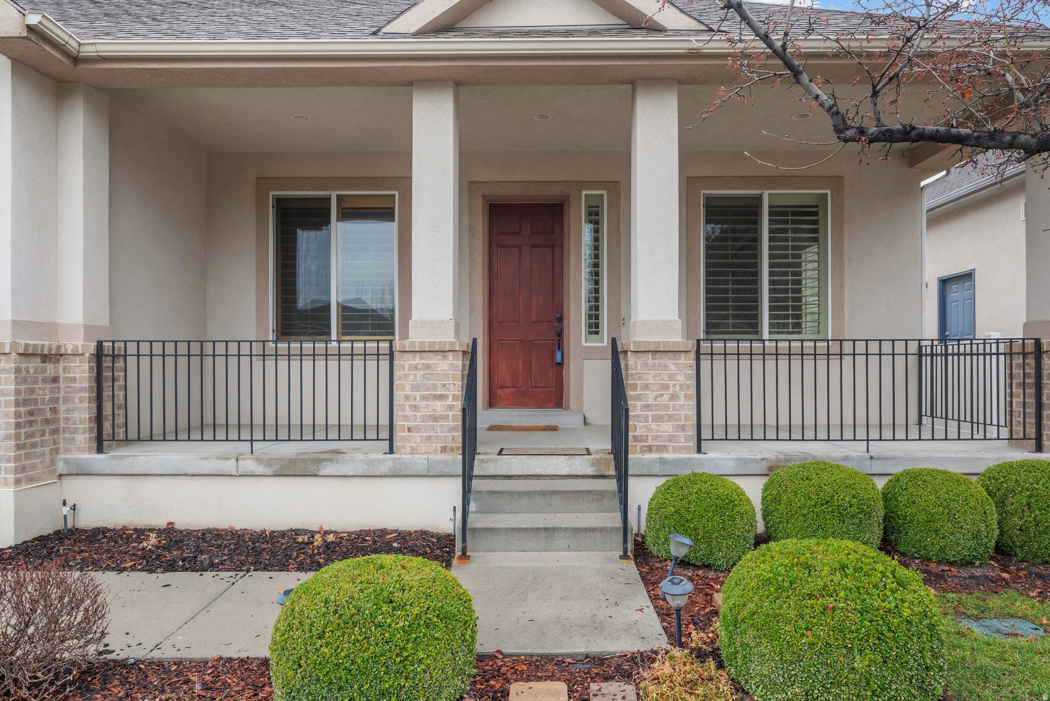 Front entry porch with brick and stucco detailing, featuring a wood-panel door, wrought iron railing, and manicured shrubbery