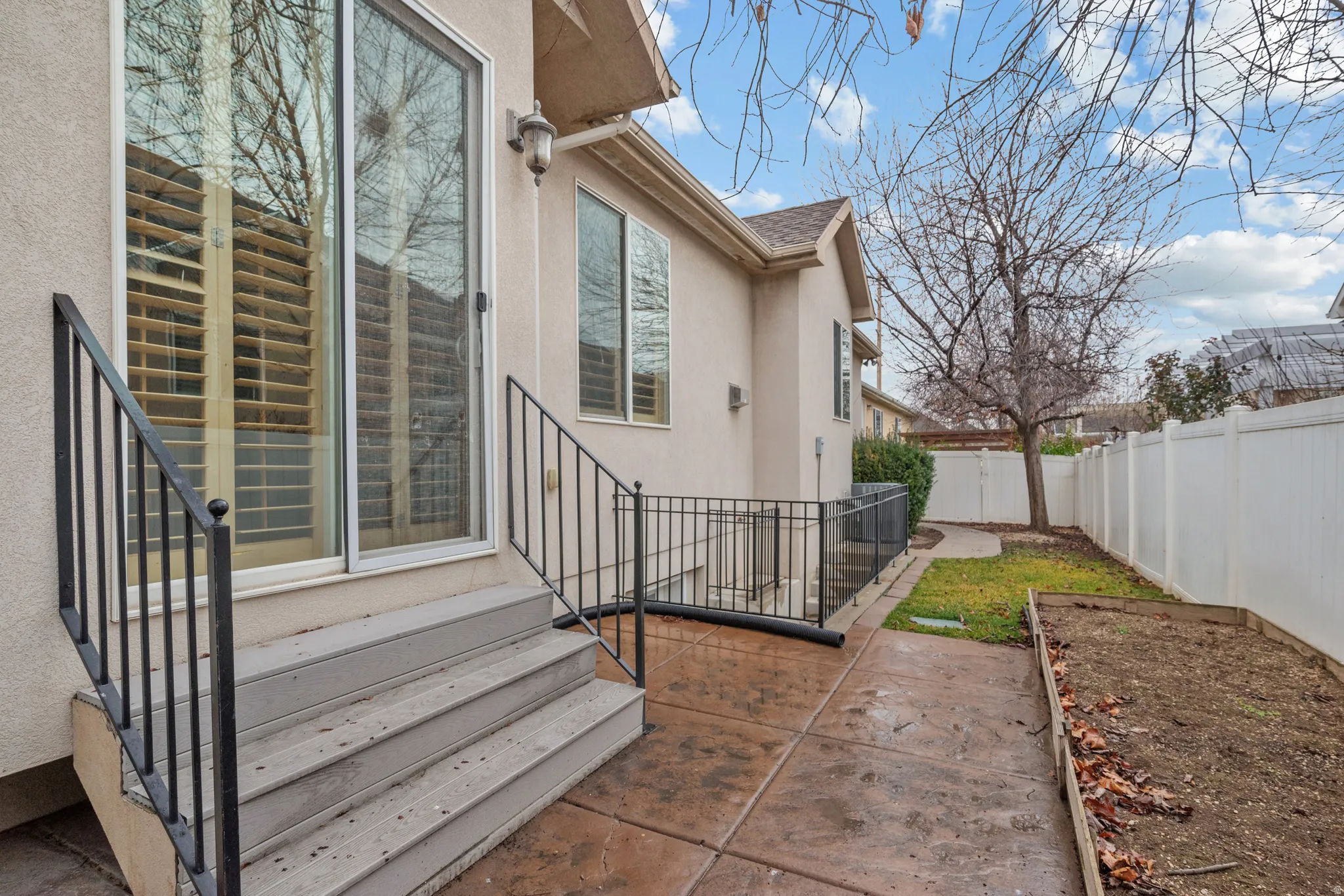 Stucco exterior featuring a sliding glass patio door, three-tier concrete steps with black metal railings, a stamped concrete patio, and an adjacent yard area with a raised garden bed and white privacy fencing