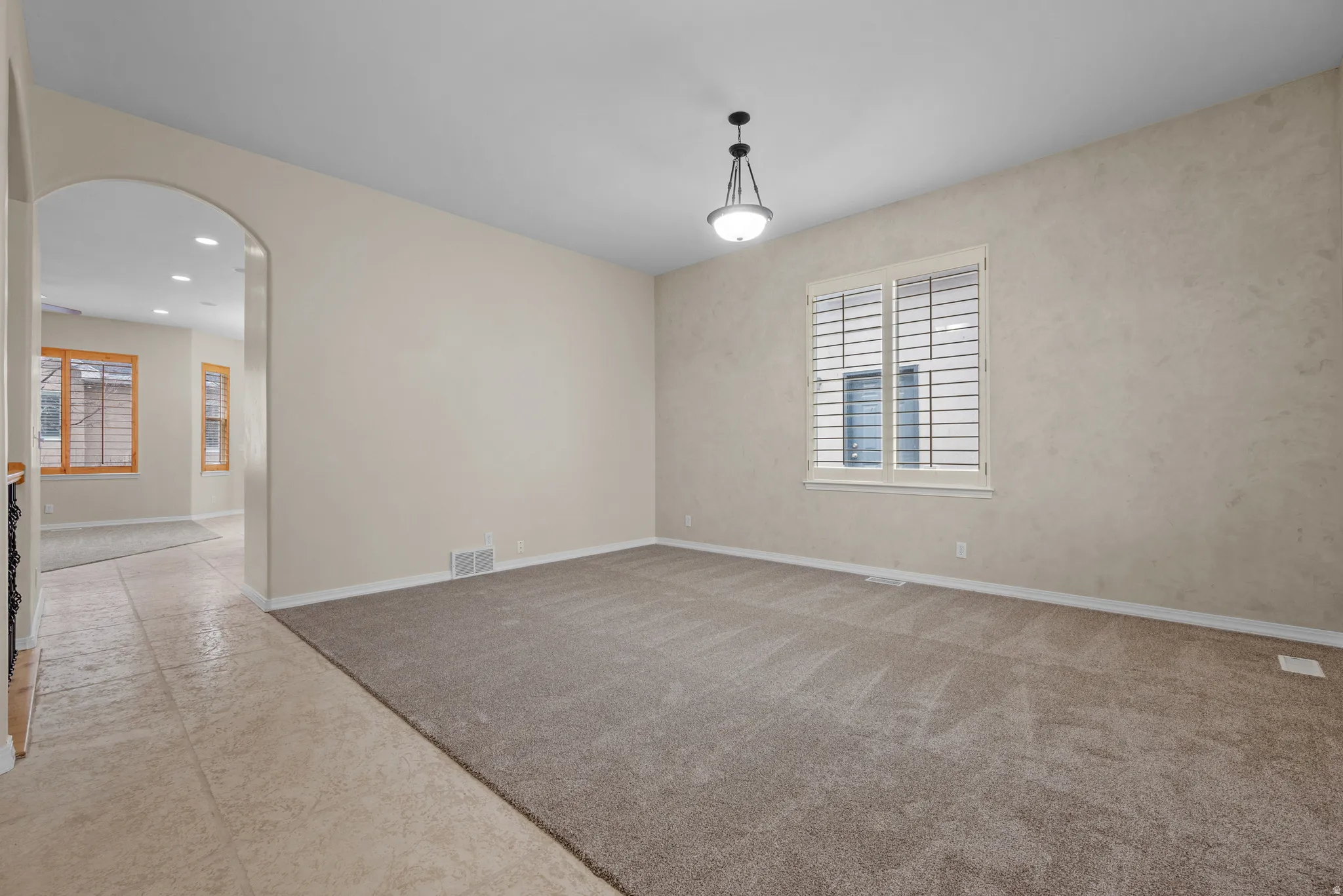 Spacious room featuring neutral carpeting, a large window with plantation shutters, and a central pendant light fixture