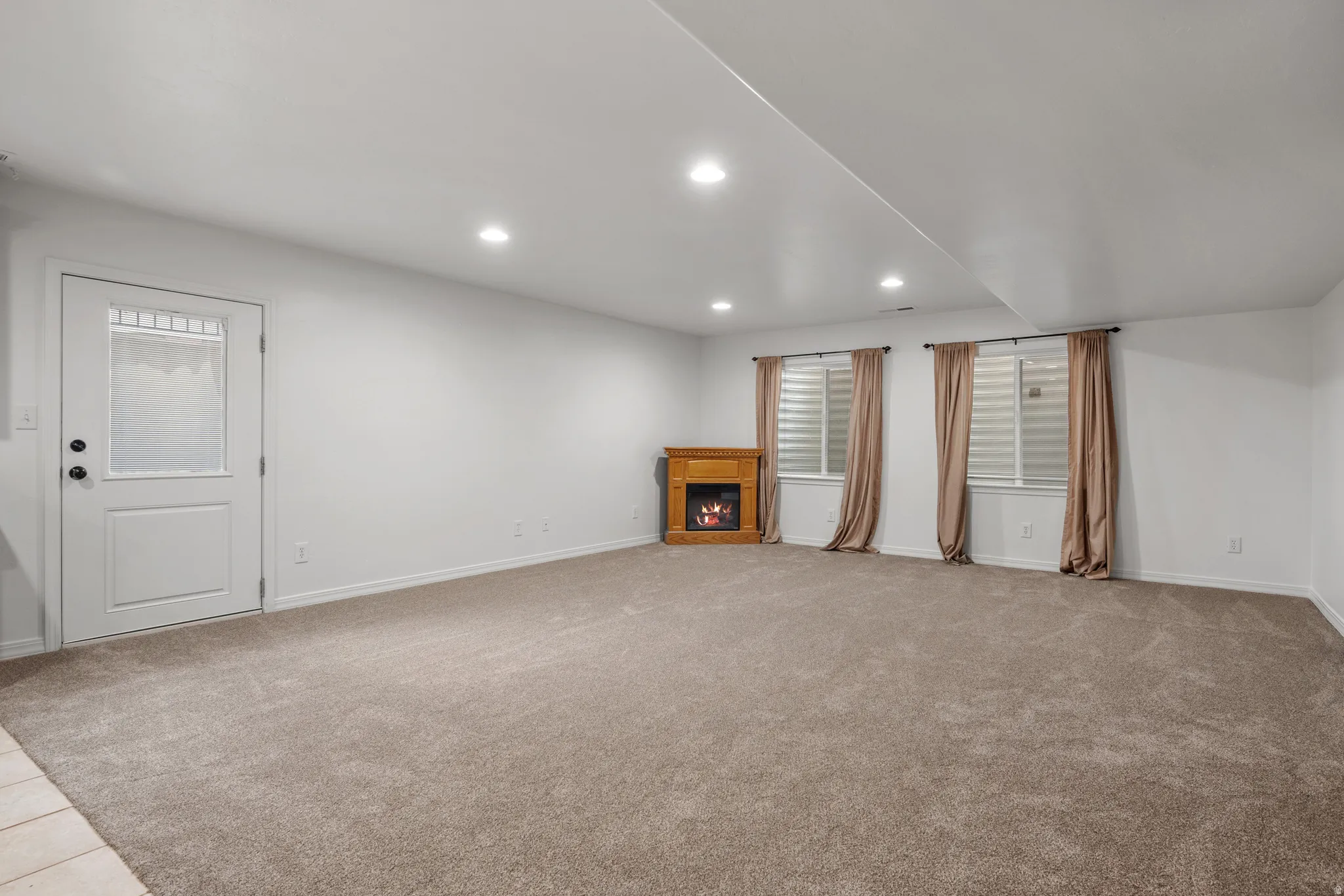 Spacious carpeted room featuring a white door with frosted glass, recessed lighting, and a wood-finish fireplace