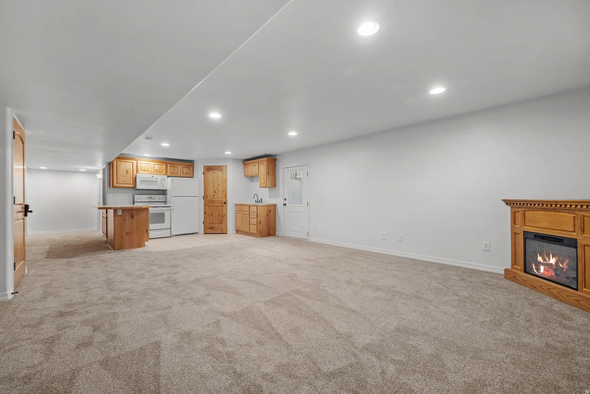 Open-concept room featuring light-toned carpet flooring, recessed lighting, a corner electric fireplace with a wood mantel, a kitchenette with wood cabinetry and white appliances, and a built-in wood island