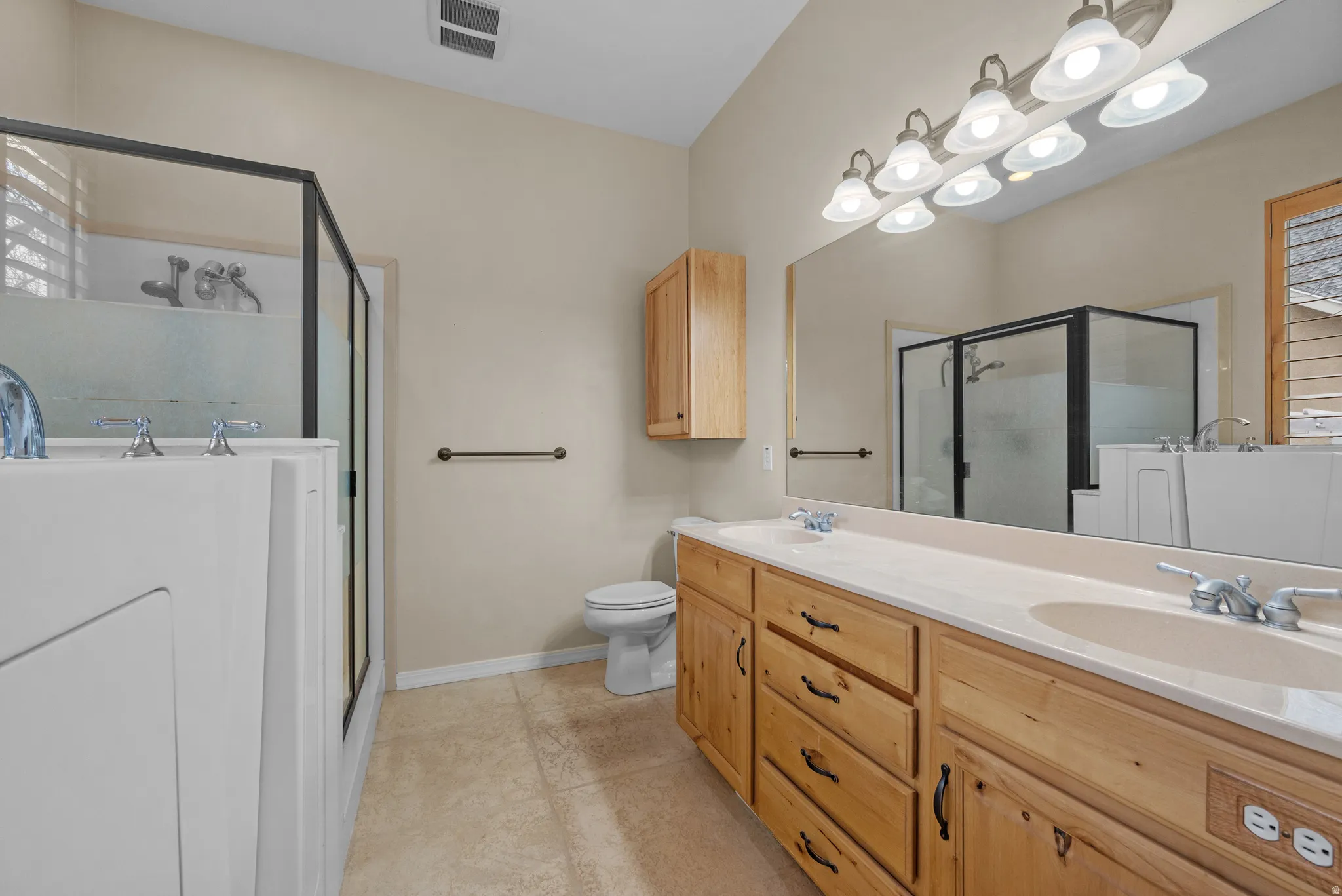 Bathroom featuring a dual vanity with light wood cabinetry and integrated sinks, a built-in tub, a separate glass-enclosed shower, and neutral tile flooring