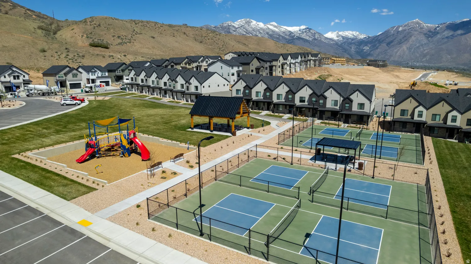 Community amenities featuring multiple pickleball courts, playground with shade structures, and a central pavilion, all surrounded by green spaces and residential buildings with mountain backdrops