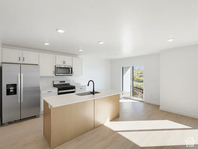 Modern kitchen featuring white cabinetry, stainless steel appliances, a light wood-finish island with integrated sink, and light wood-finish flooring