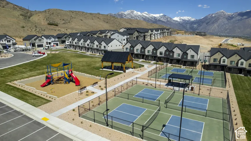 Community amenities featuring multiple pickleball courts, playground with shade structures, and a central pavilion, all surrounded by green spaces and residential buildings with mountain backdrops