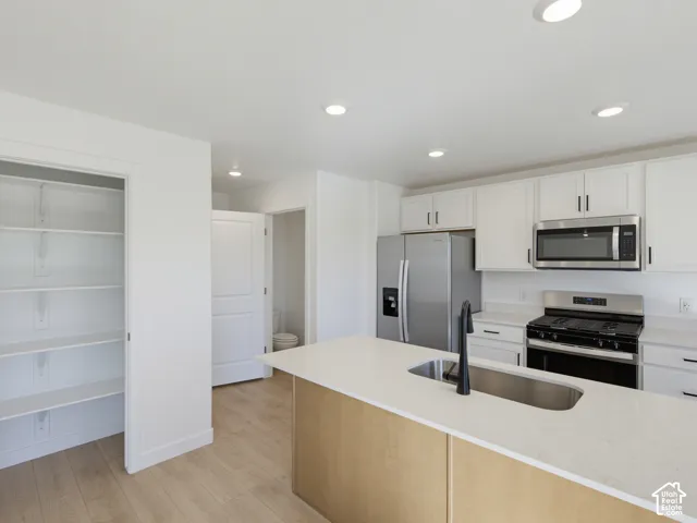 Kitchen featuring a light-toned island with an undermount sink, stainless steel appliances, white cabinetry, and wood-finish flooring