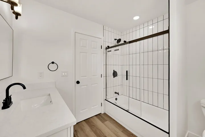 Bathroom featuring a white subway tile shower surround with black hardware, a shower/tub combination with a sliding glass door, a vanity with an integrated sink and light-toned countertop, black faucet, and wood-finish flooring