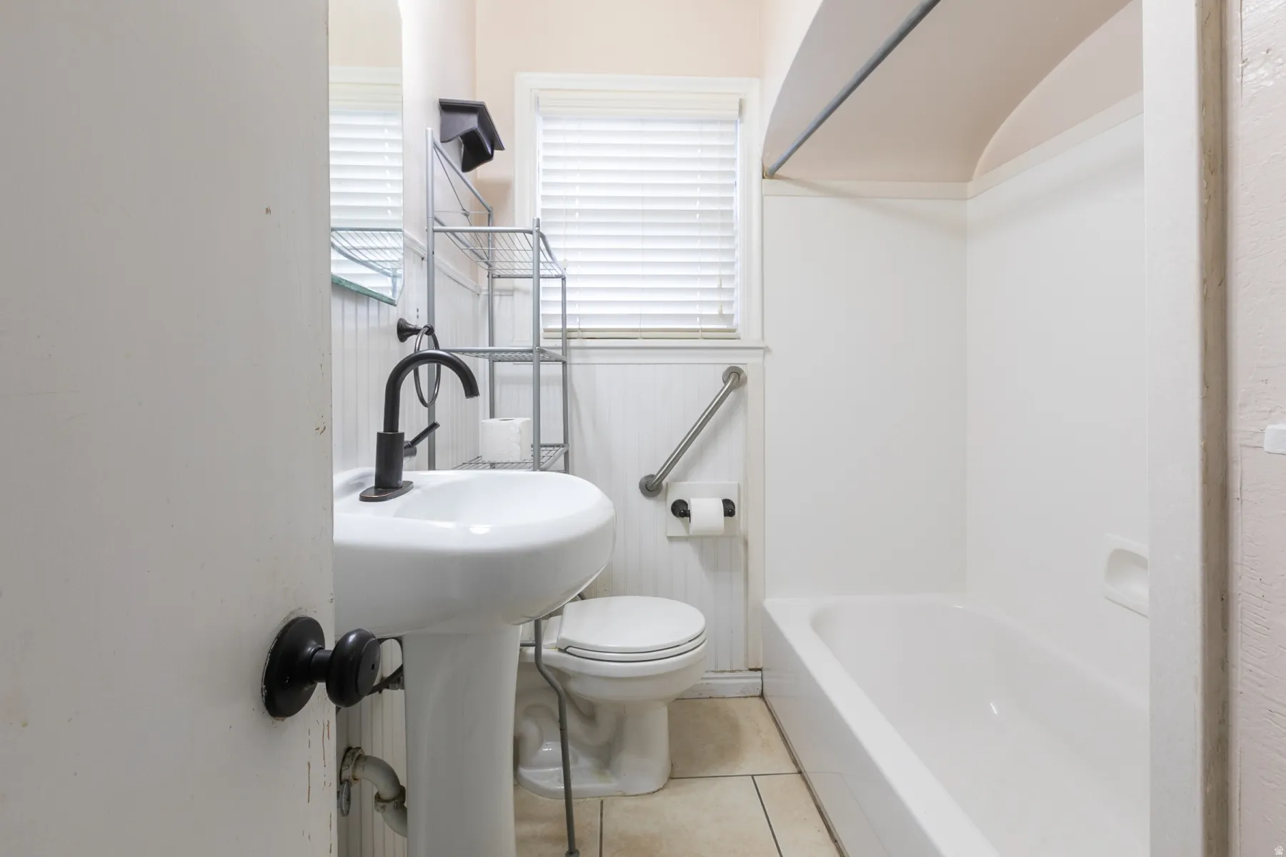 Bathroom featuring a white pedestal sink with a matte black faucet, ceramic tile flooring, beadboard wainscoting, and a white bathtub with an integrated grab bar
